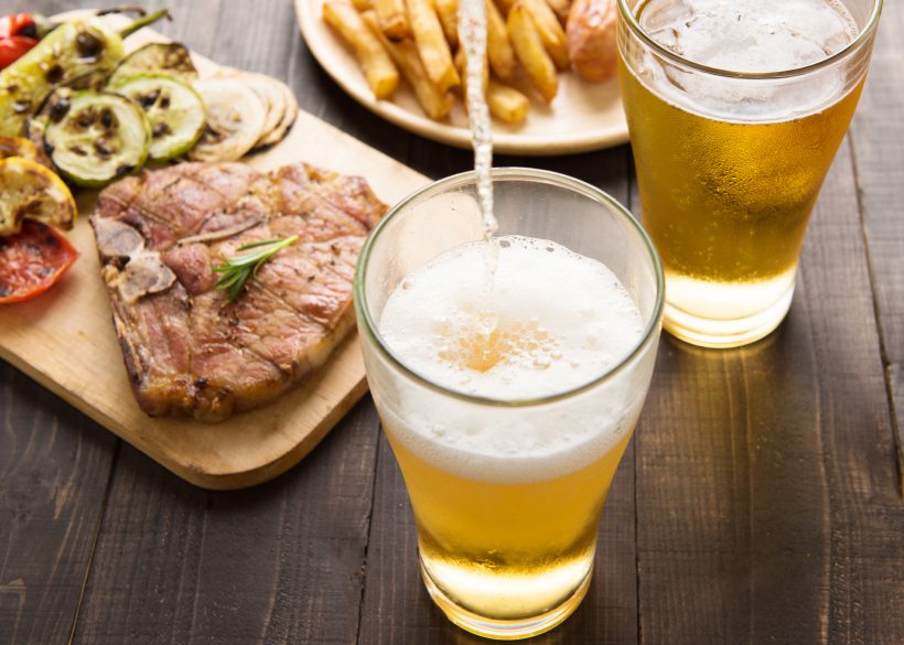 Beer being poured into glass with gourmet steak and french fries on wooden background.